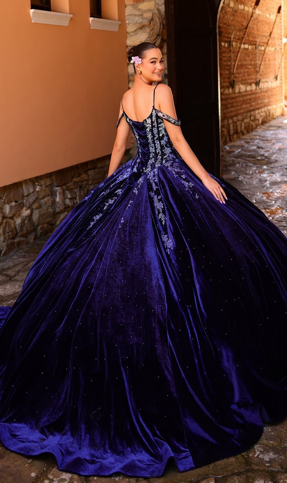 A woman in the Quinceanera Dress 54316 by Amarra, an off-the-shoulder ball gown with ombre lace embroidery, stands outdoors on a stone pathway, smiling over her shoulder.